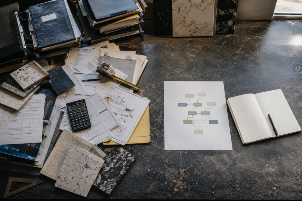 Stone samples, equipment documents, production notes, and a simple flow plan on a fabricator owner's worktable, representing growth complexity and profitable system design.