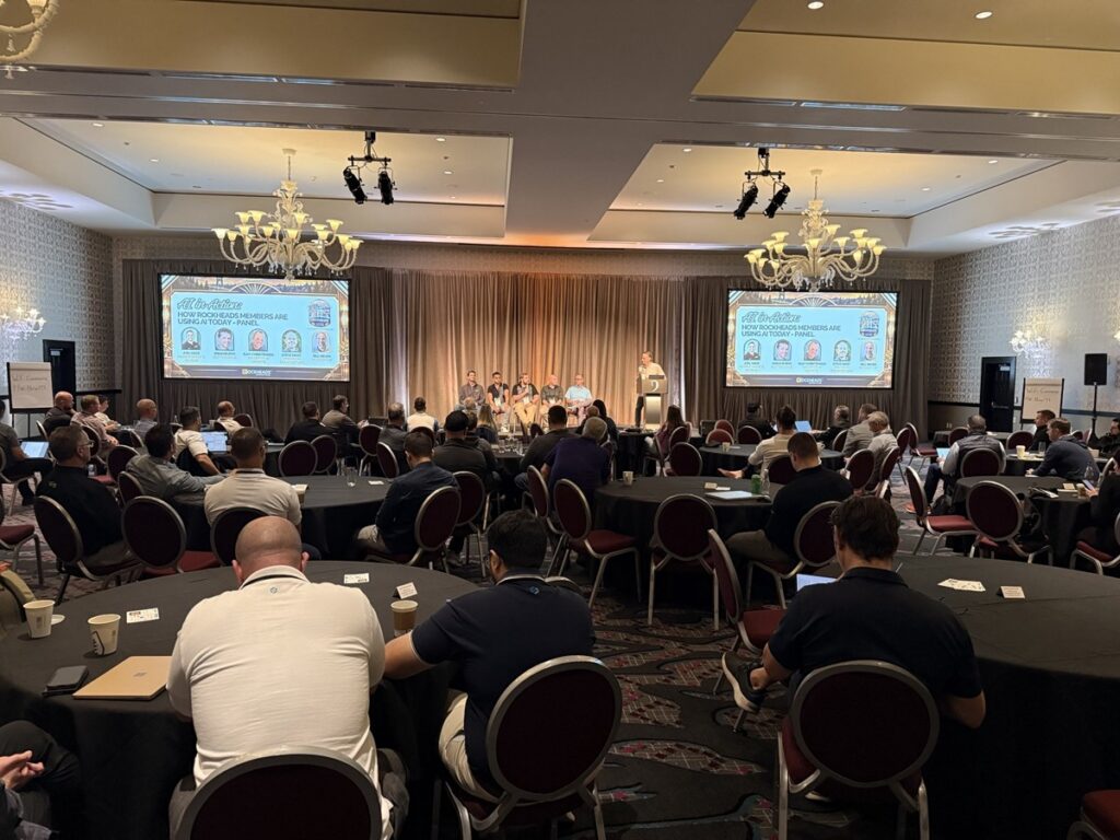 Wide view of the Rockheads Portland presentation room, with attendees seated at round tables facing panelists and two large screens.